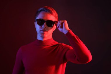 Güneş gözlüğü takan yakışıklı adam karanlık arka plana karşı neon ışıklarla bakıyor.