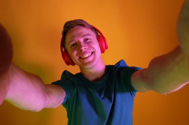 Kulaklık takan gülümseyen adam turuncu arka planda selfie çekiyor.