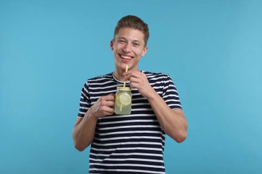 Mason kavanozundan açık mavi arka planda limonata içen gülümseyen adam. Tazeleyici içecek.