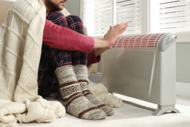 Evde ısıtıcının yanında elleri ısıtan bir adam var.