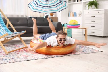 Güneş gözlüklü mutlu adam ve şişme yüzüklü paletler evde yerde eğleniyor. Denizde tatilin hayalini kuruyorum.