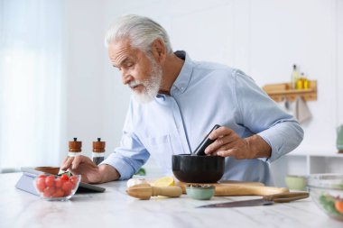 Mutfaktaki beyaz mermer masada yemek pişirirken tablet kullanan yaşlı adam.