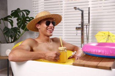 Güneş gözlüklü ve hasır şapkalı mutlu adam evdeki küvette şişme yüzüklü mason kavanozundan meyve suyu içiyor. Denizde tatilin hayalini kuruyorum.