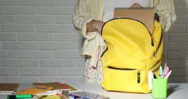 Masadaki sarı çantaya farklı kırtasiye malzemesi koyan kadın, yakın plan.