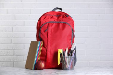 Kırmızı sırt çantası, farklı kırtasiye malzemesi ve beyaz tuğla duvara karşı hafif mermer masa üzerinde elma.