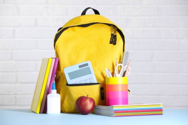 Sarı sırt çantası, farklı kırtasiye malzemesi ve beyaz tuğla duvara karşı açık mavi masada elma.