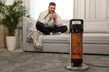 Evde elektrikli ısıtıcının yanında bir bardak sıcak içeceği olan adam seçici bir şekilde odaklanmış.