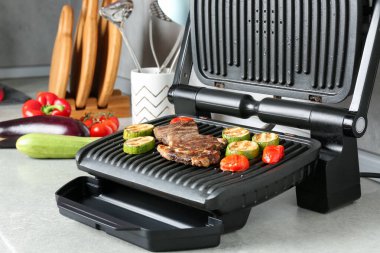 Açık gri masada biftek ve sebzelerle elektrikli ızgara, yakın plan.