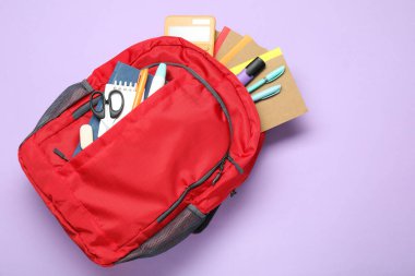 Menekşe arkaplan üzerinde farklı kırtasiye malzemesi olan kırmızı sırt çantası, üst görünüm