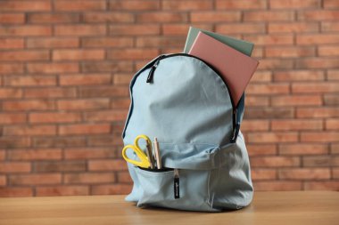 Kırmızı tuğla duvara karşı ahşap masanın üzerinde farklı kırtasiye malzemesi olan açık mavi sırt çantası.