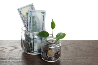 Glass jars with dollars, sprout and coins on wooden table against white background