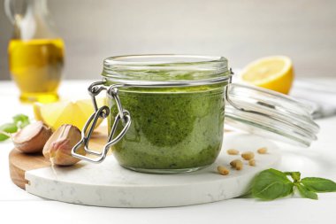 Cam kavanozda lezzetli yeşil pesto ve beyaz ahşap masada malzemeler, yakın plan.