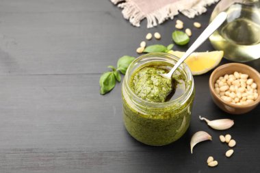 Kavanozda kaşıkla taze pesto sosu ve siyah ahşap masada malzemeler, yakın plan. Metin için boşluk