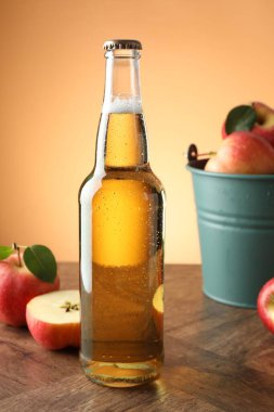 Delicious cider in glass bottle and apples on wooden table against orange gradient background