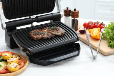 Mutfaktaki beyaz mermer masada lezzetli et, sebze ve baharatlarla elektrikli ızgara, yakın plan.