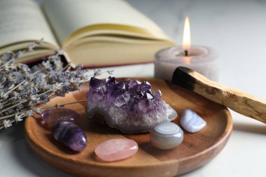 Farklı güzel değerli taşlar, palo santo, kitap, lavanta çiçekleri ve beyaz masada yanan mum, yakın plan.
