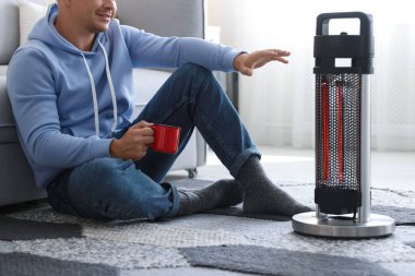 Evinde elektrikli ısıtıcının yanında bir bardak içkisi olan genç adam, yakın çekim.