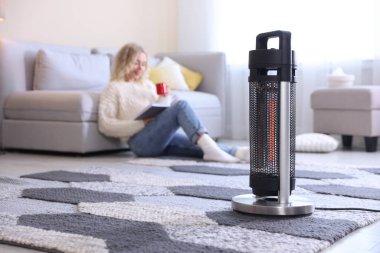 Elinde bir bardak içki ve kitapla evde elektrikli ısıtıcının yanında duran genç kadın, seçici bir şekilde odaklanmış.