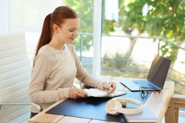 Not defterine yazan ve içerideki ahşap masada dizüstü bilgisayar kullanan genç bir kız. Ev işyeri