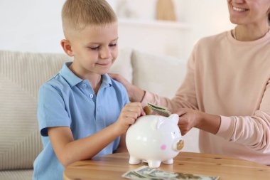 Anne ve oğlu, kapalı ahşap masadaki domuz kumbarasına cep harçlığı koyuyorlar.