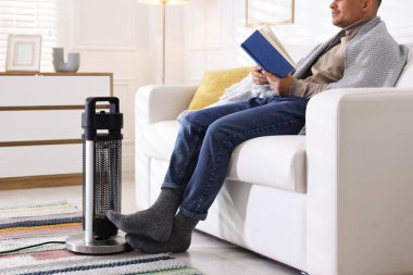 Evde elektrikli ısıtıcının yanında kitap okuyan genç adam, yakın çekim.