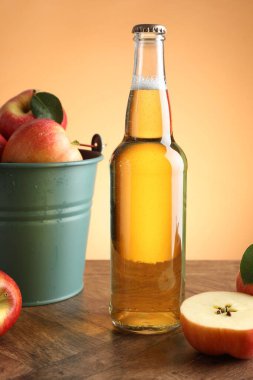 Delicious cider in glass bottle and apples on wooden table against orange gradient background