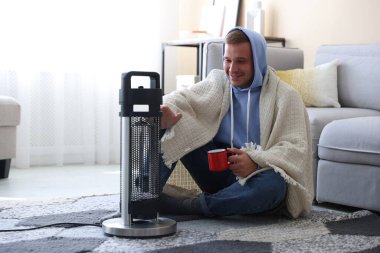 Evinde elektrikli ısıtıcının yanında bir bardak içkisi olan genç adam.