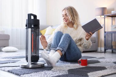 Elinde bir bardak içki ve kitapla evde elektrikli ısıtıcının yanında oturan genç bir kadın.