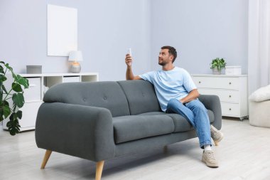 Evdeki kanepede uzaktan kumandayla klima kullanan bir adam.