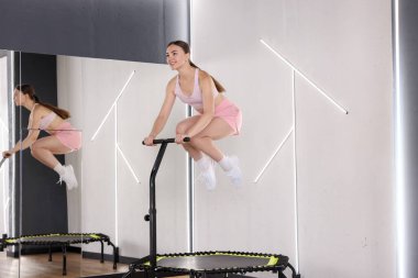 Spor salonundaki aynanın yanındaki trambolinde zıplayan gülümseyen kadın.