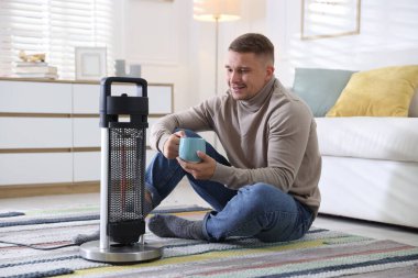 Evinde elektrikli ısıtıcının yanında bir bardak içkisi olan genç adam.