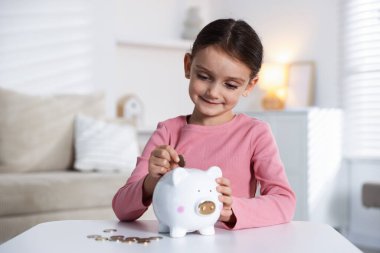 Küçük kız, içerideki beyaz masadaki kumbaraya cep harçlığı koyuyor.