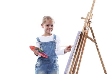 Meslek seçimi. Beyaz arka planda ressammış gibi davranan palet boyalı ve sehpanın yanındaki fırçalı küçük kız.