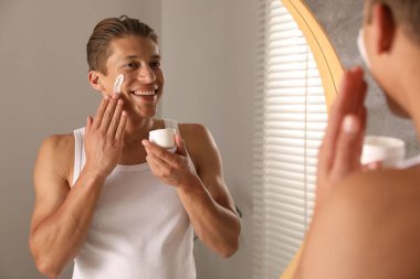 Yakışıklı genç adam banyoda aynanın yanında yüzüne krem sürüyor.