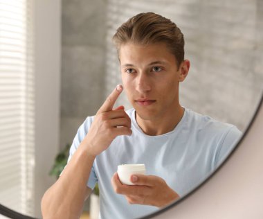 Yakışıklı genç adam evde aynanın yanında yüzüne krem sürüyor.