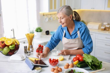 Mutfaktaki beyaz mermer masada yemek pişirirken tablet kullanan yaşlı kadın.