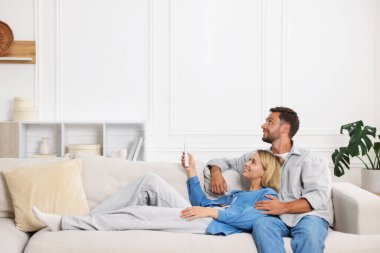 Evdeki kanepede klima kontrol etmek için uzaktan kumanda kullanan çift. Metin için boşluk