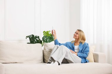 Evdeki koltukta uzaktan kumandayla klima kullanan bir kadın.