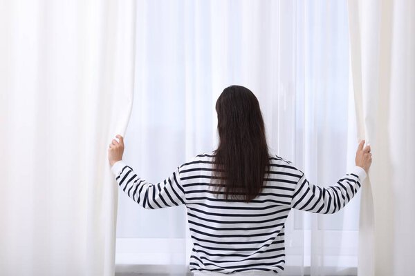 Woman opening window curtains at home, back view