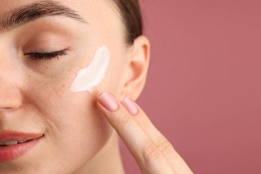 Güzel bir kadın pembe arka planda yüzüne krem sürüyor, yakın plan. Metin için boşluk