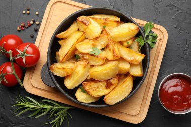 Kızartma tavasında maydanozlu fırında patates, biberiye, ketçap ve domatesler koyu dokuma sehpasında.