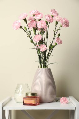 Beautiful carnation flowers in vase and candles on white stand near beige wall indoors