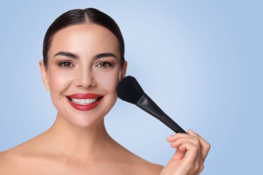 Güzel bir kadın açık mavi arka planda fırçayla makyaj yapıyor.