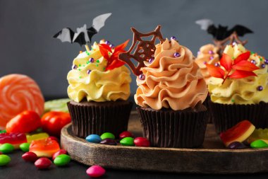 Gri arka planda, üzerinde yarasa süsü, çikolata parçacıkları, çikolata ağı ve renkli şekerler olan enfes kapkekler. Cadılar Bayramı