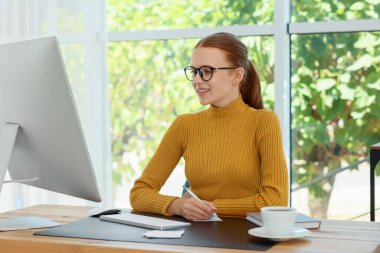 Genç kız bilgisayarını kullanıyor ve içerideki ahşap masada deftere yazı yazıyor. Ev işyeri