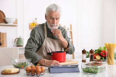 Mutfaktaki beyaz mermer masada yemek pişirirken tablet kullanan yaşlı adam.