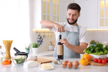 Gülümseyen adam mutfakta yemek pişirirken şarap şişesini açıyor.