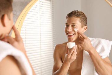 Yakışıklı genç adam banyoda aynanın yanında yüzüne krem sürüyor.