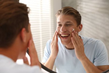 Yakışıklı genç adam evde aynanın yanında yüzüne krem sürüyor.