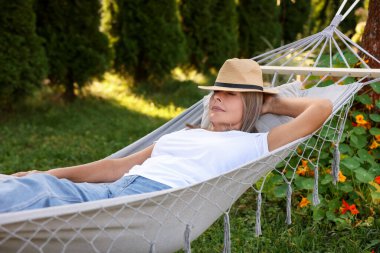 Hasır şapkalı yaşlı kadın dışarıda hamakta dinleniyor.
